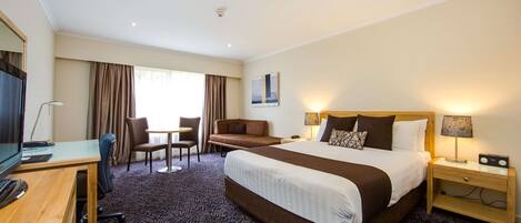 A hotel room with a large bed, a desk with a computer, a chair, a small table, and a TV.