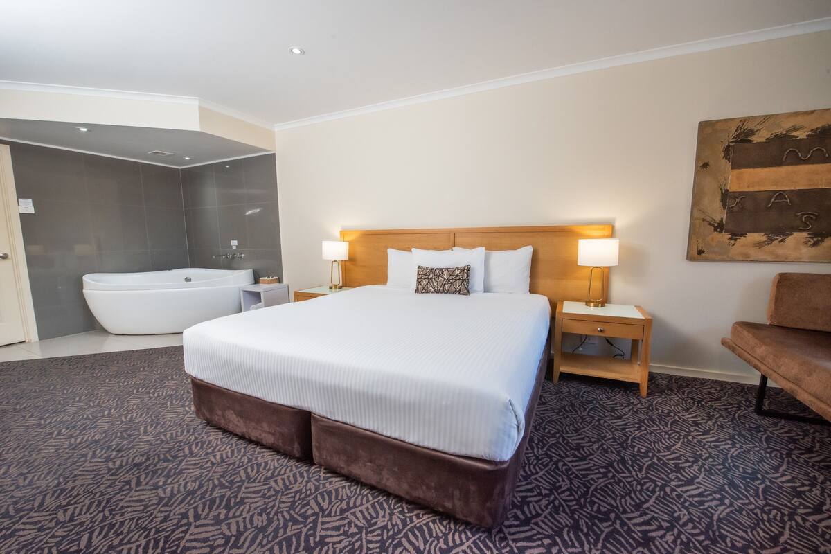 A hotel room with a large bed, a wooden headboard, two bedside lamps, a nightstand, a brown bench, a large bathtub, and a wall-mounted artwork.