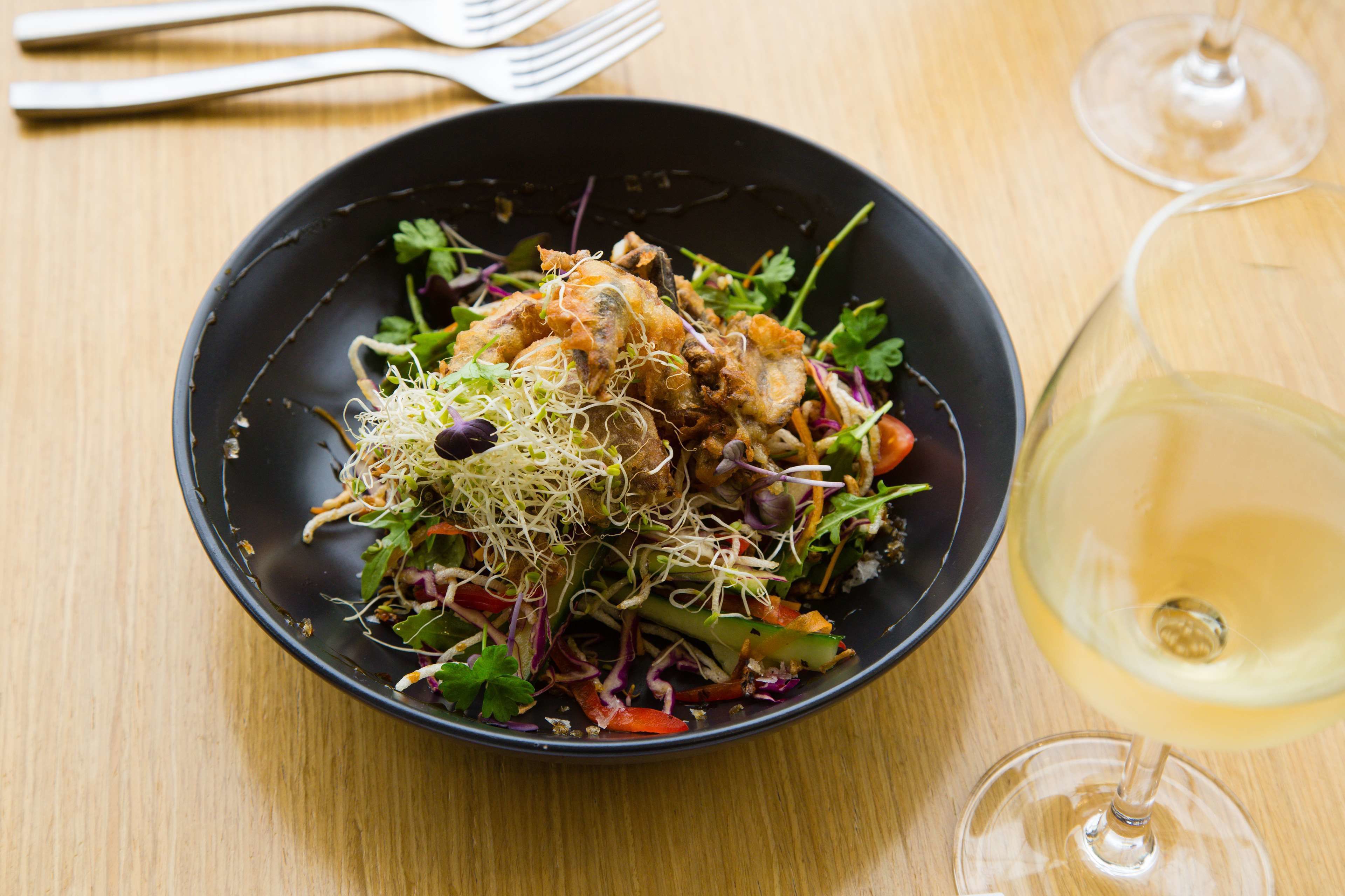 A black plate with a salad, a glass of white wine, and a fork on a wooden table.
