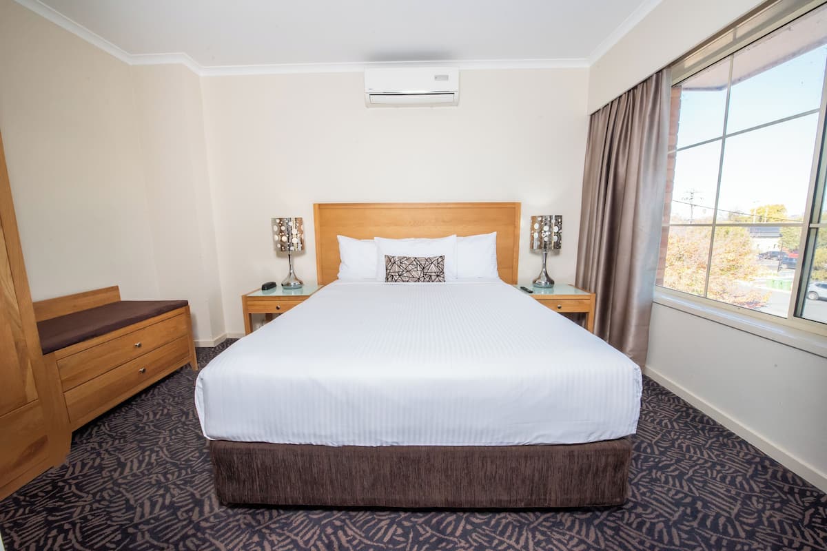 A hotel room with a large bed, wooden headboard, two bedside tables with lamps, a wooden bench, and a window with curtains.