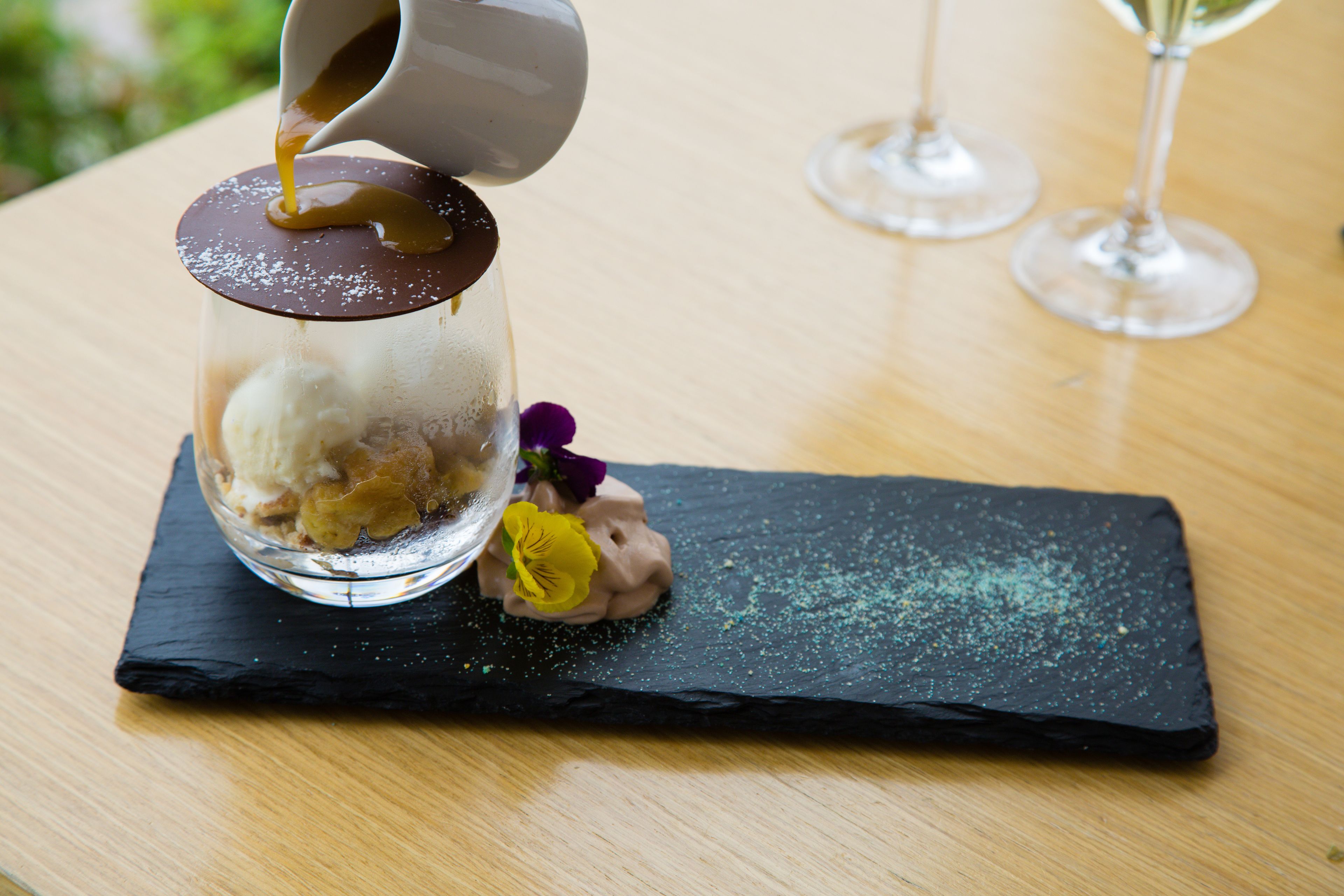 Un postre en vaso con una cúpula de chocolate, al que se vierte salsa de caramelo, sobre una losa negra.