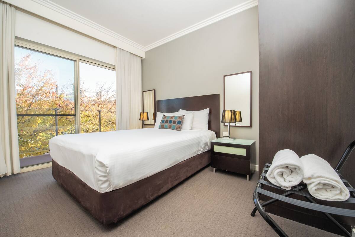 A hotel room with a large bed, a nightstand, a lamp, and a view of autumn trees through the window.
