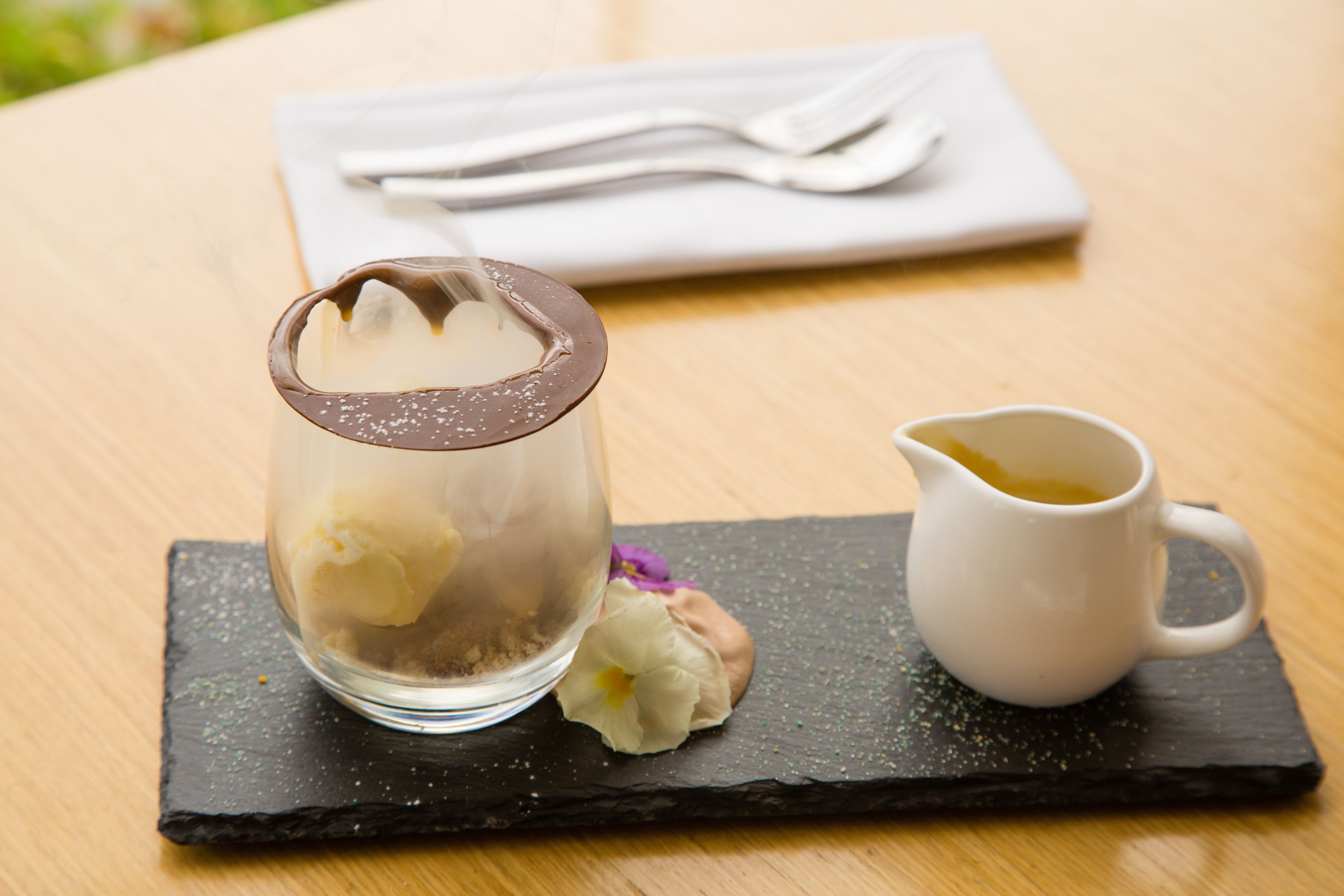 A dessert in a glass container with a chocolate lid, a white pitcher, and a folded napkin with utensils on a wooden table.