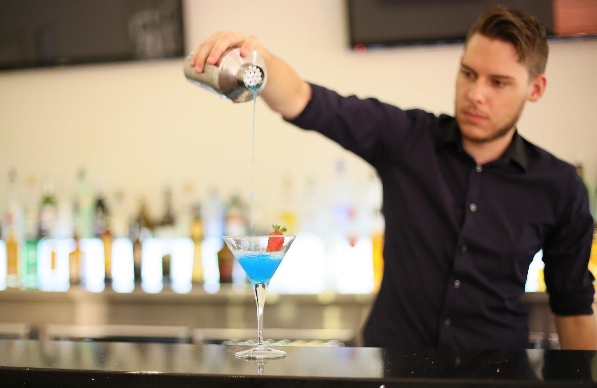 A bartender pouring a blue cocktail into a martini glass with a strawberry garnish.