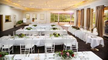 Un salón de banquetes con varias mesas redondas dispuestas para el servicio de comidas, cada una con manteles blancos y centros de mesa florales.