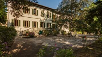 A two-story building with a courtyard, featuring a stone pathway, outdoor seating, and lush greenery.