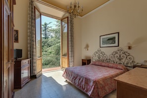 A bedroom with a bed, a desk, a television, and a balcony with a view of trees and a building. - Giotto Park Hotel (Vaglia)