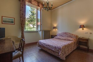 A bedroom with a bed, a desk, and a chair. There is a window with curtains, a chandelier, and wall-mounted lamps. - Giotto Park Hotel (Vaglia)