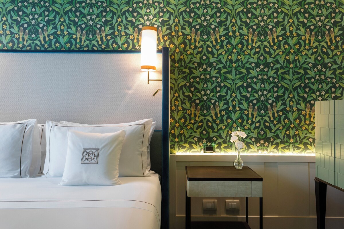 A hotel room with a bed, pillows, a nightstand, a vase with flowers, and a wall with a floral pattern.