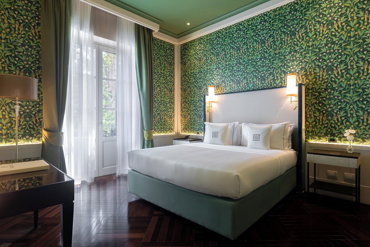 A hotel room with a large bed, a desk, and a chair. There is a window with curtains, a nightstand with a lamp, and a wall with a floral pattern.