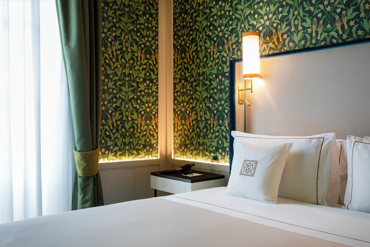 A hotel room with a bed, a nightstand, a lamp, and a window with curtains.