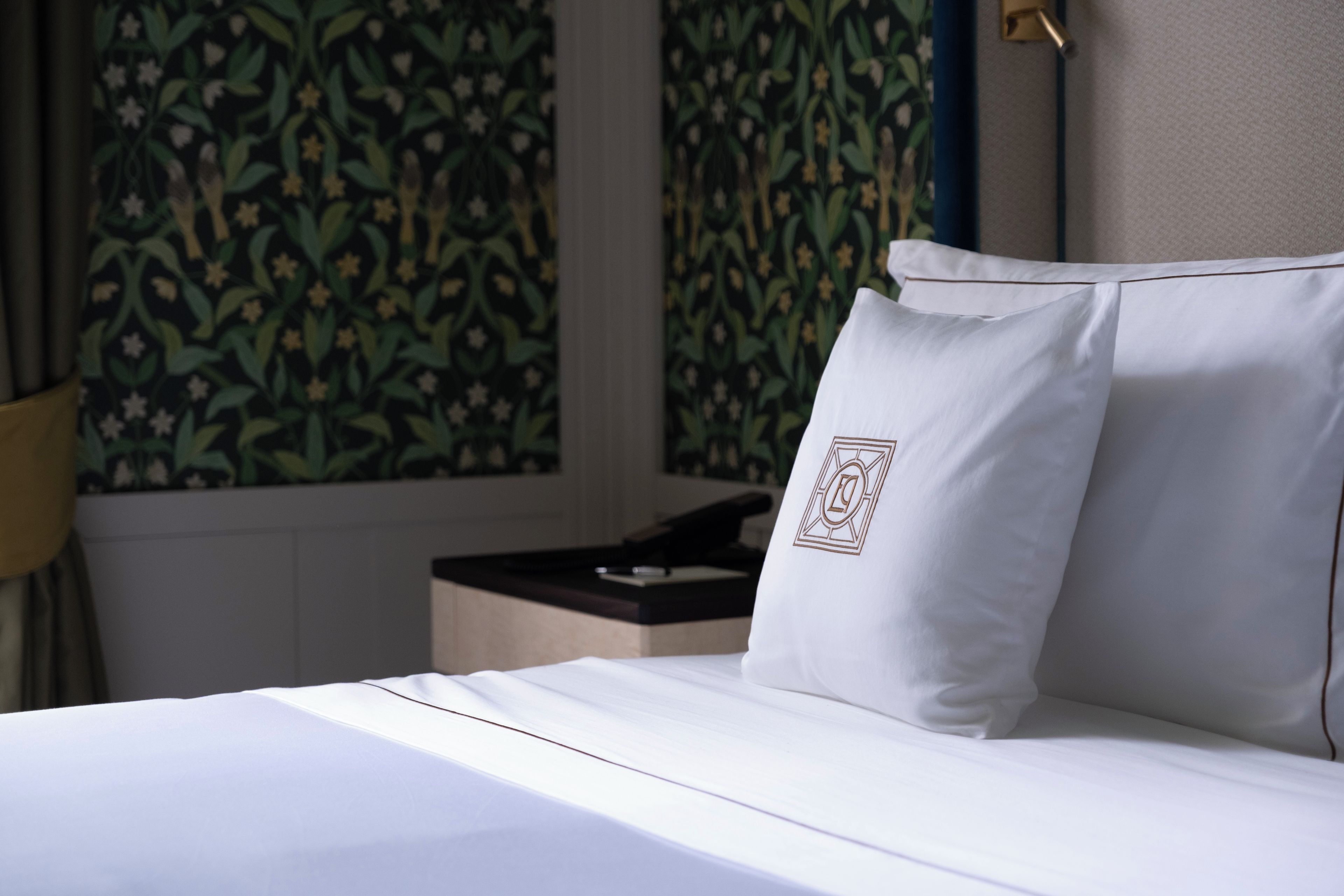 A neatly made bed with white linens and pillows, a bedside table with a remote control, and a wall with a floral pattern.