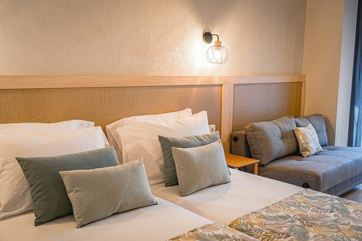 A hotel room with two beds, a sofa, a nightstand, and a wall-mounted light fixture.