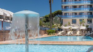 En swimmingpool med en fontän, solstolar och en byggnad med balkonger i bakgrunden.