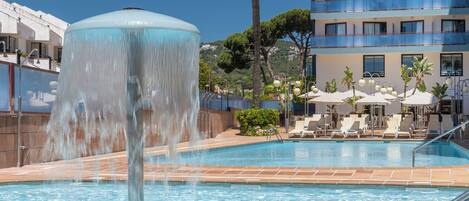 A swimming pool with a water fountain, lounge chairs, and a building with balconies in the background.