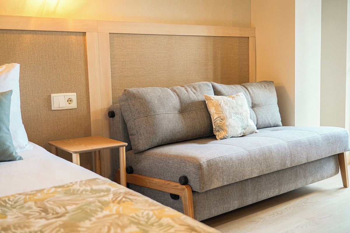 A modern hotel room with a grey sofa, a wooden nightstand, and a bed with a patterned bedspread.