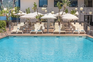 A poolside area with lounge chairs and umbrellas, a clear pool, and a building with windows and balconies in the background. - htop Jadhe (Calella)