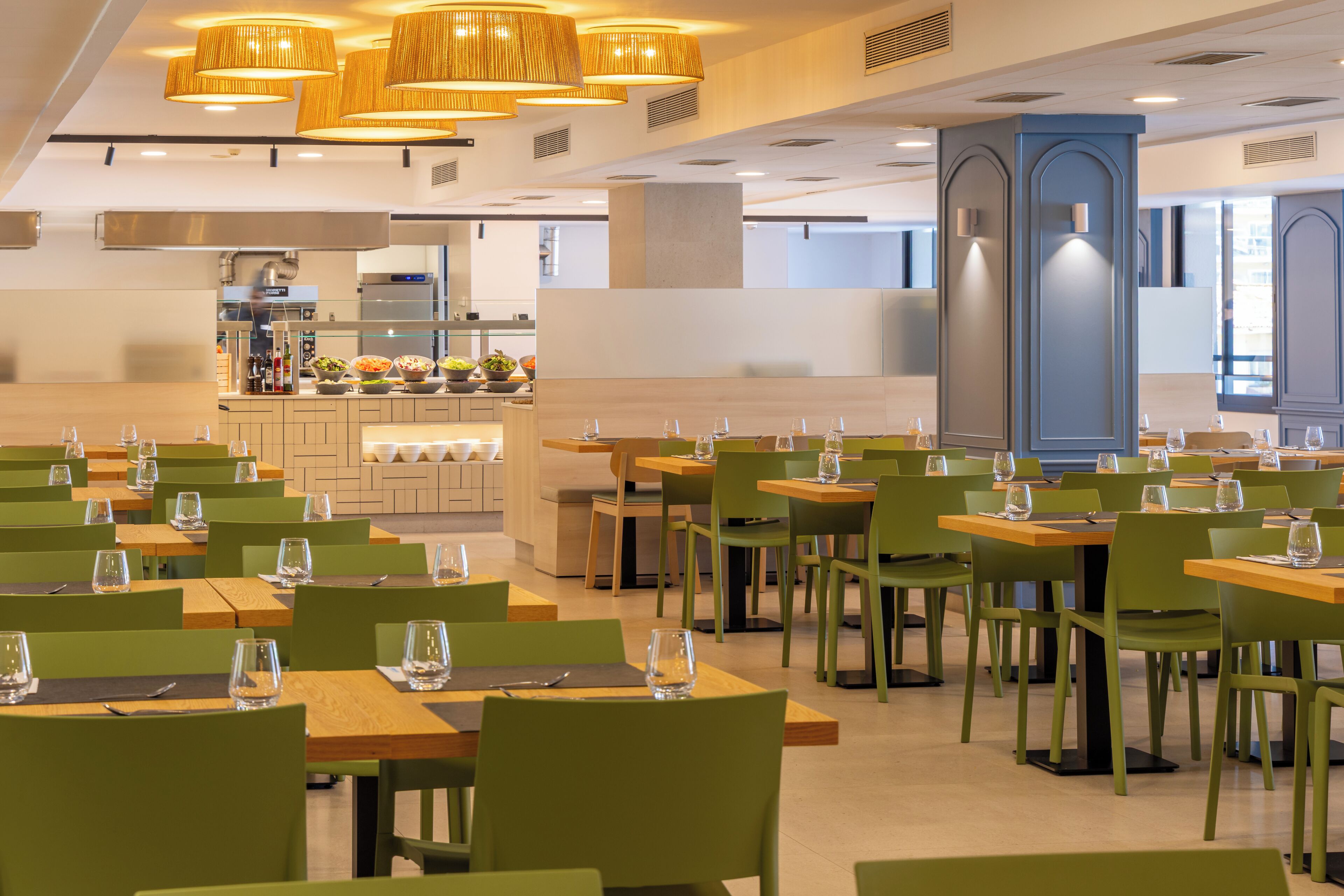 Un restaurante con varias mesas y sillas, un área de cocina al fondo e iluminación colgante.