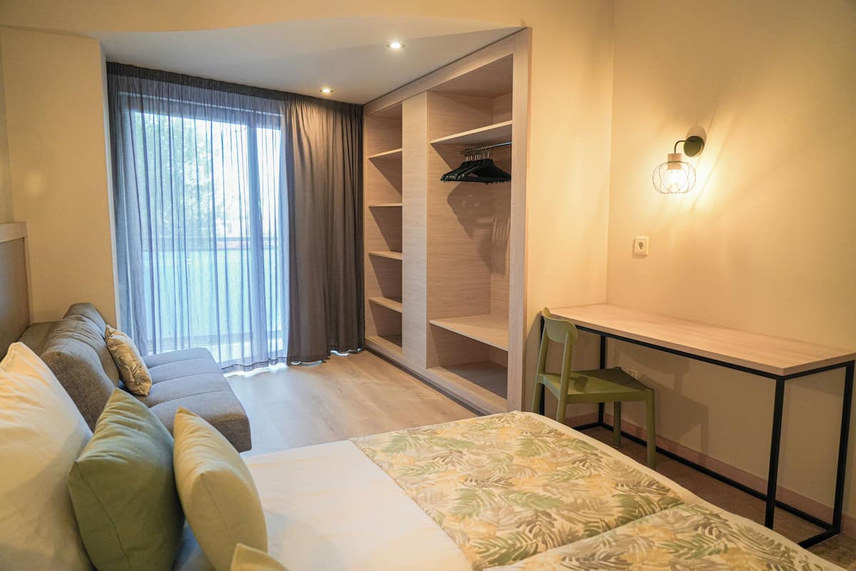 A bedroom with a bed, a green chair, a desk, a wardrobe, and a window with curtains.