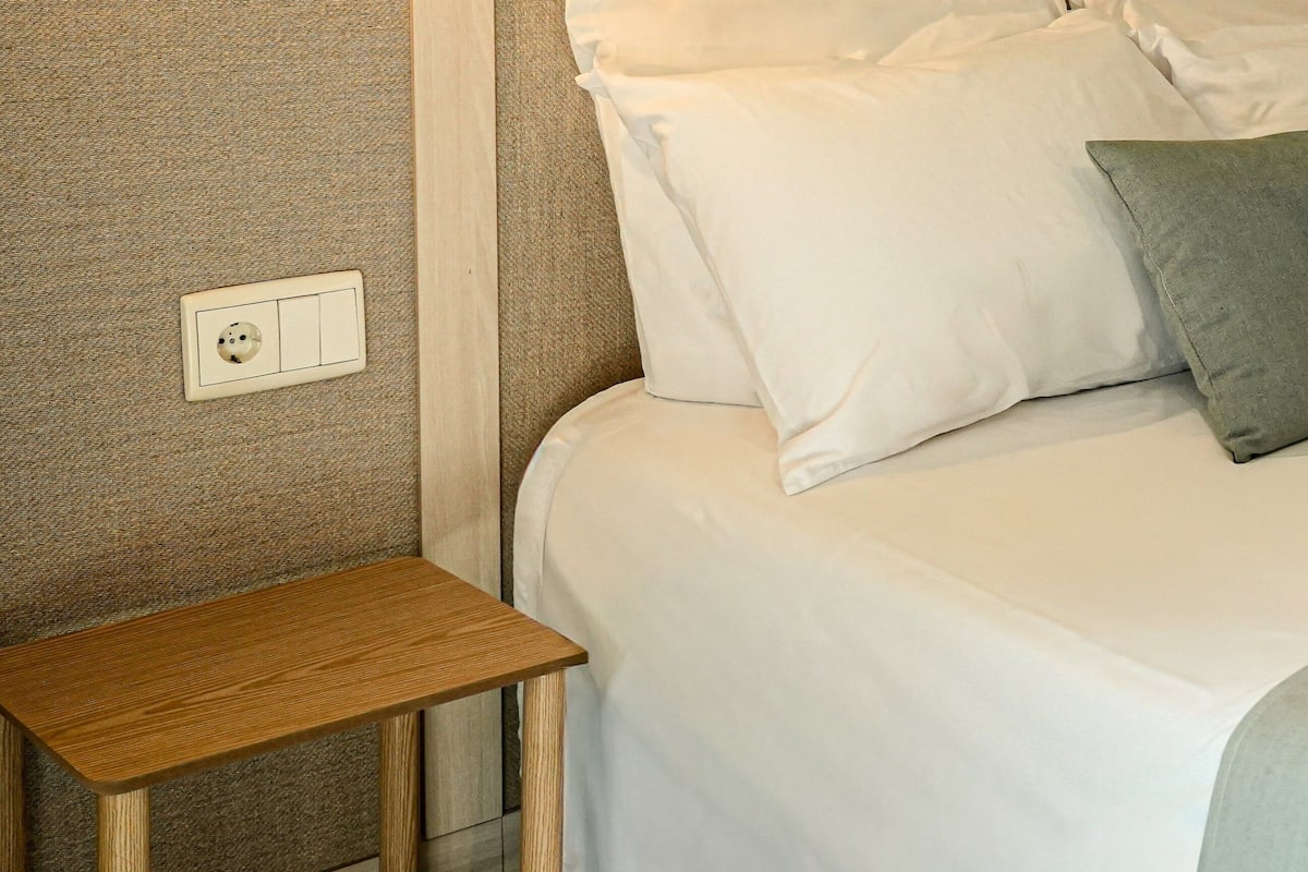 A neatly made bed with white bedding and two pillows, one plain and one with a leaf pattern. A small wooden nightstand is positioned to the left of the bed.