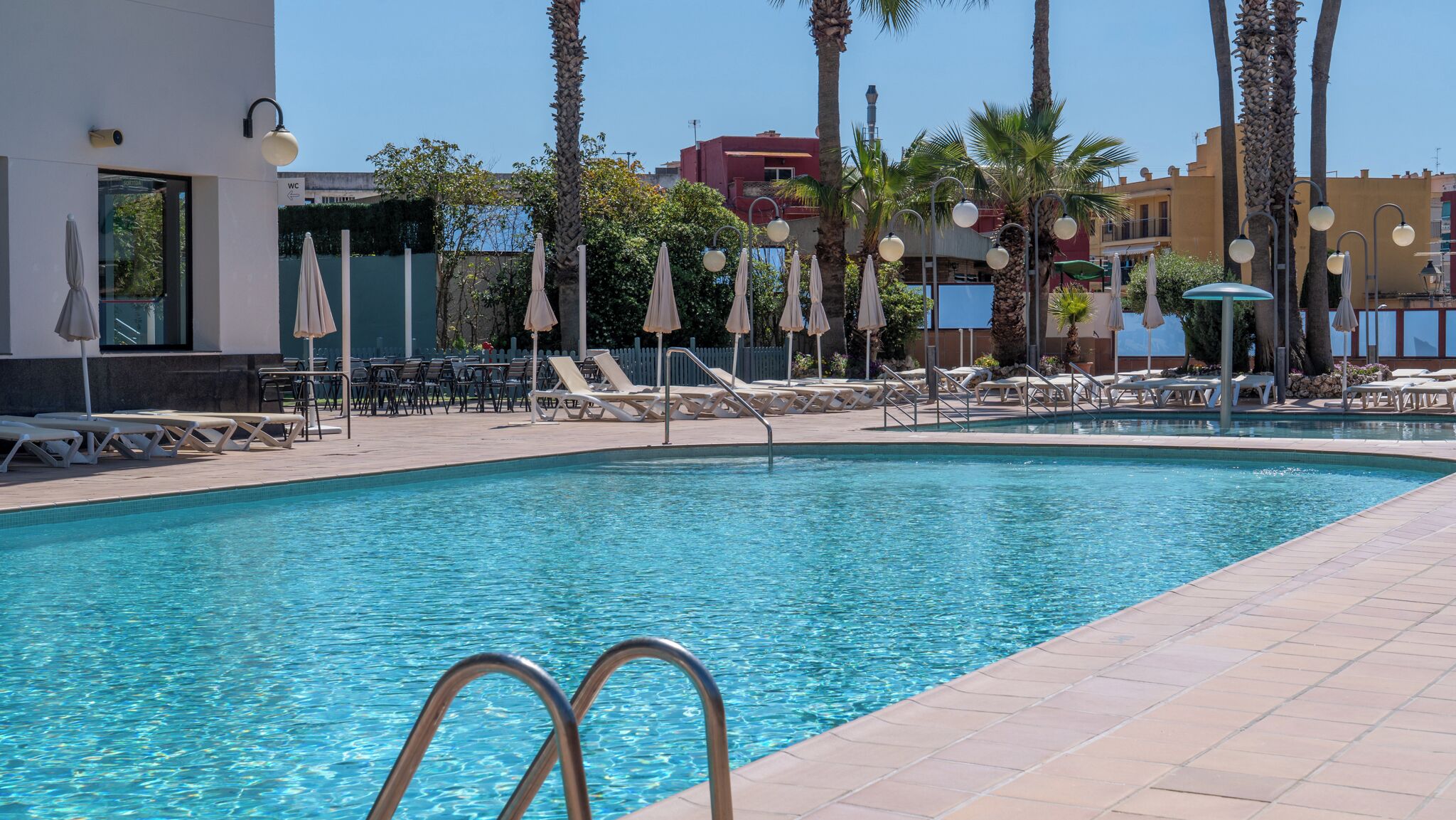 A swimming pool with lounge chairs and umbrellas, surrounded by palm trees and buildings.