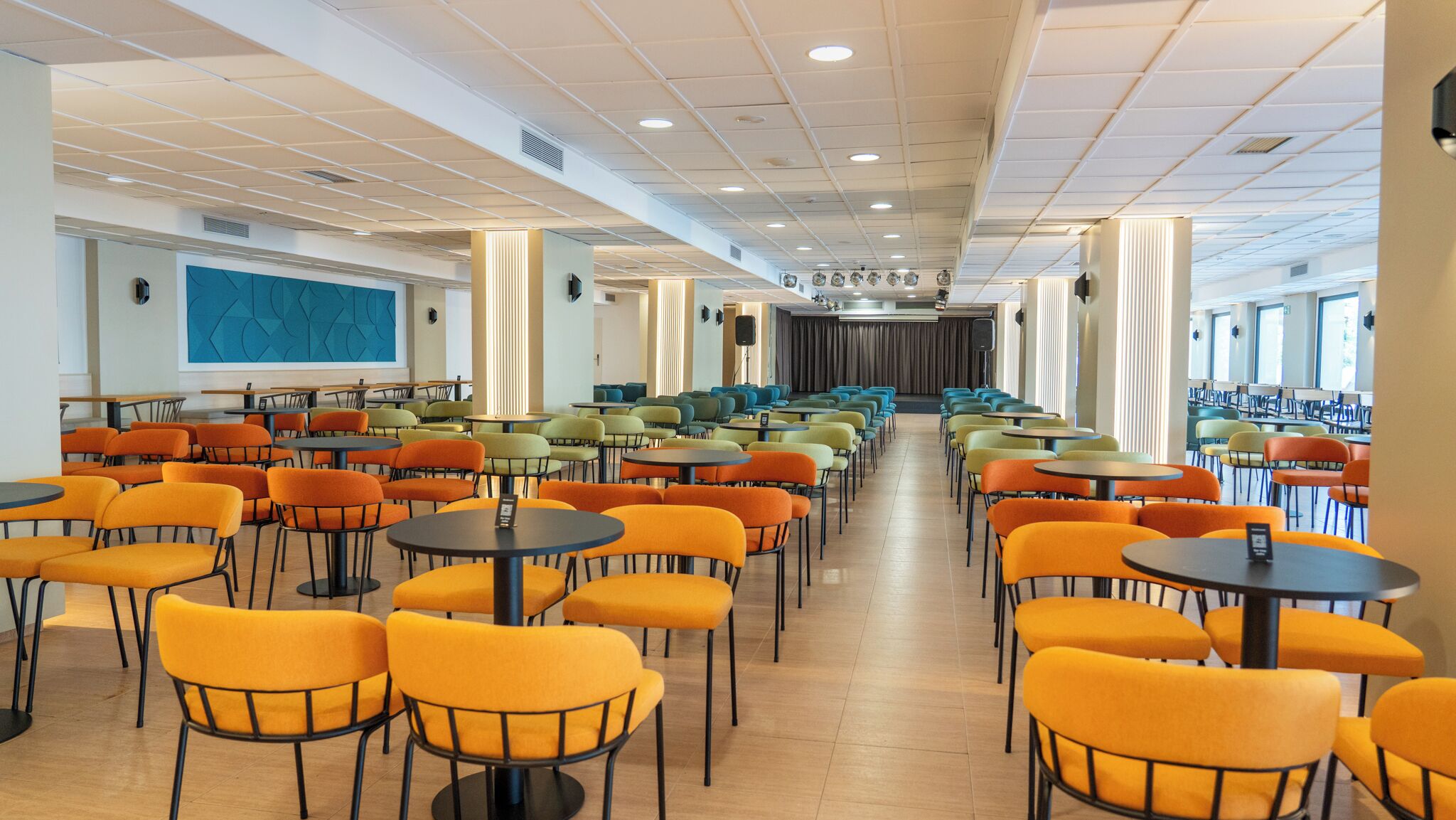 Un comedor amplio con filas de mesas y sillas, una pantalla grande a la izquierda y un escenario al fondo.