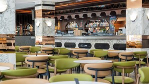 Un restaurant amb diverses taules i caires, una zona de bar amb diferents ampolles, i una paret de marbre.