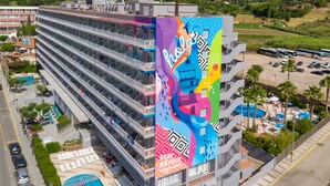 Hotel de diverses plantes amb balcons, una piscina i un mural colorit que anuncia l'hotel.