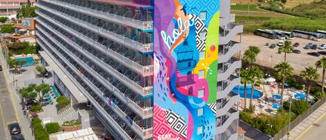 Un hotel de varios pisos con balcones, piscina y un mural colorido que anuncia el hotel.