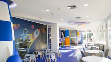 Zona infantil con murales coloridos, estructuras de juego y mesas y sillas dispuestas para actividades.