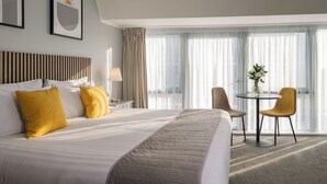Habitación de hotel con una cama grande, una mesita de noche con l ámpara, una pequeña mesa redonda con sillas y vistas a la ciudad a través de grandes ventanales.
