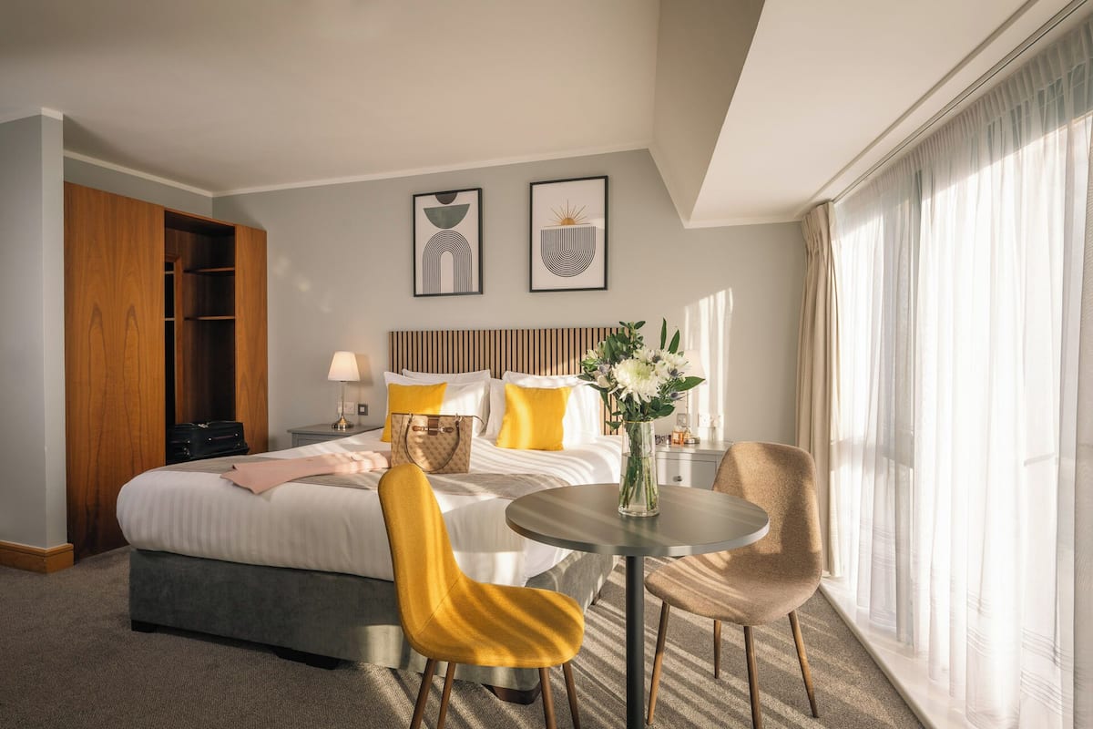 Habitación de hotel con cama, mesita de noche, una silla, una pequeña mesa y una ventana con cortinas.