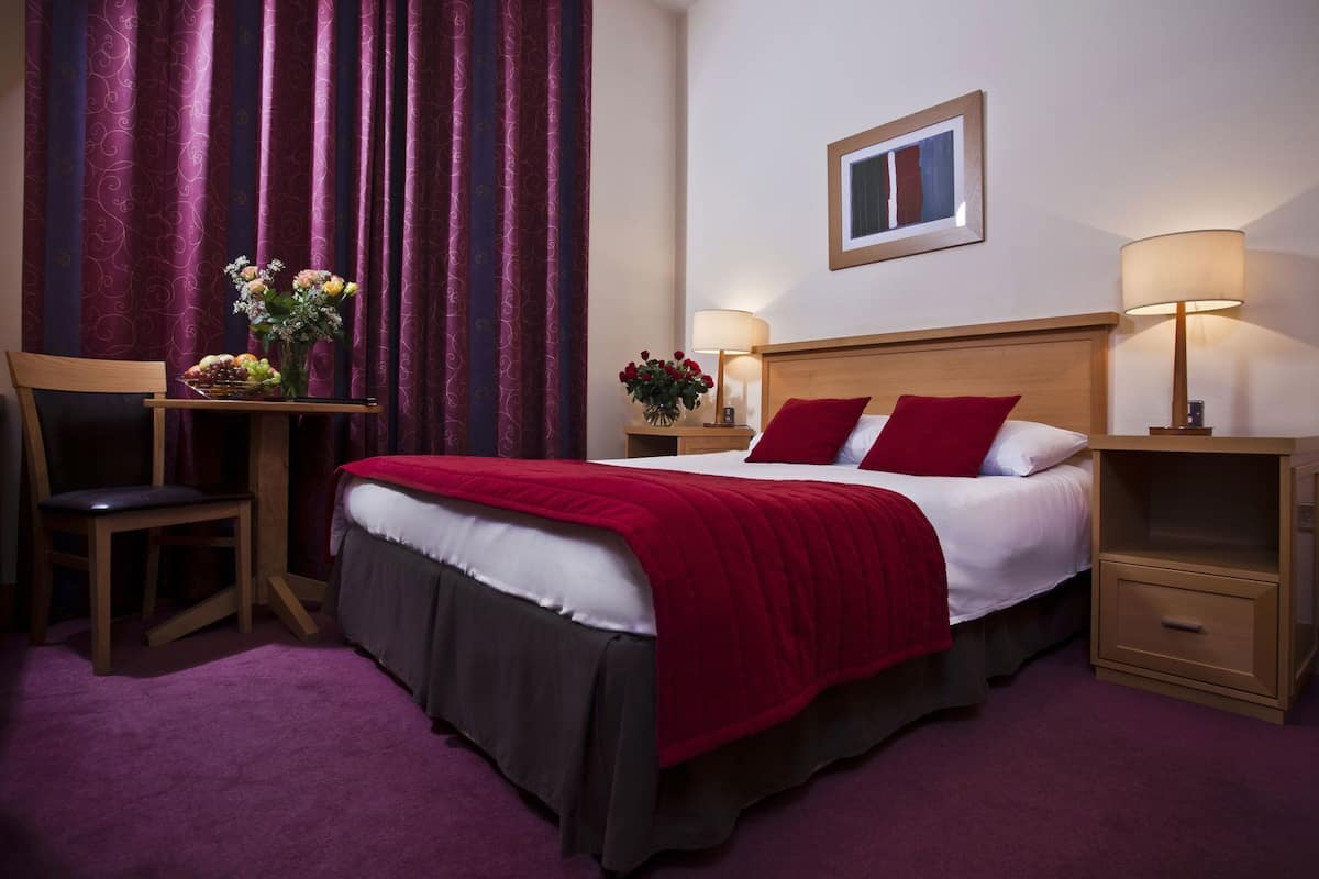 Habitación de hotel con cama, dos mesitas de noche, una silla, un escritorio con un florero y una ventana con cortinas.