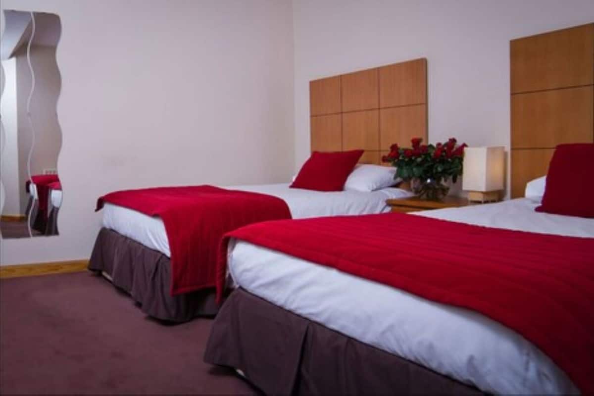 Habitación de hotel con dos camas, cada una con una manta roja y sábanas blancas, un cabecero de madera, una mesita de noche con una lámpara y un espejo.