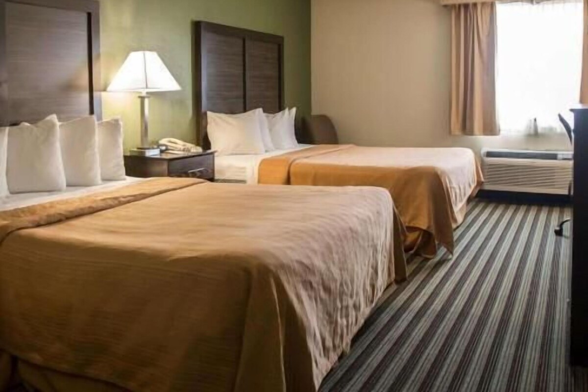 A hotel room with two beds, a nightstand with a lamp, a desk with a telephone, and a window with curtains.
