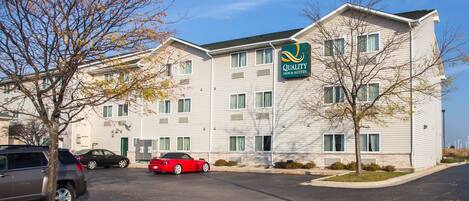 Il s’agit d’un hôtel à plusieurs étages, reconnaissable à son enseigne indiquant qu’il fait partie de la chaîne Quality Inn & Suites. Plusieurs voitures sont garées dans l’allée, dont une voiture de sport rouge.