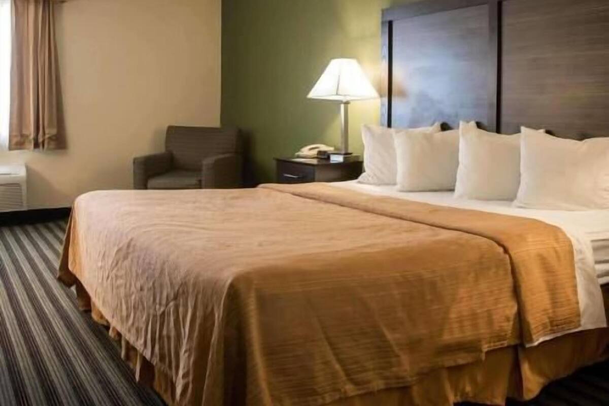 A hotel room with a large bed, a chair, a nightstand with a lamp, and a window with curtains.