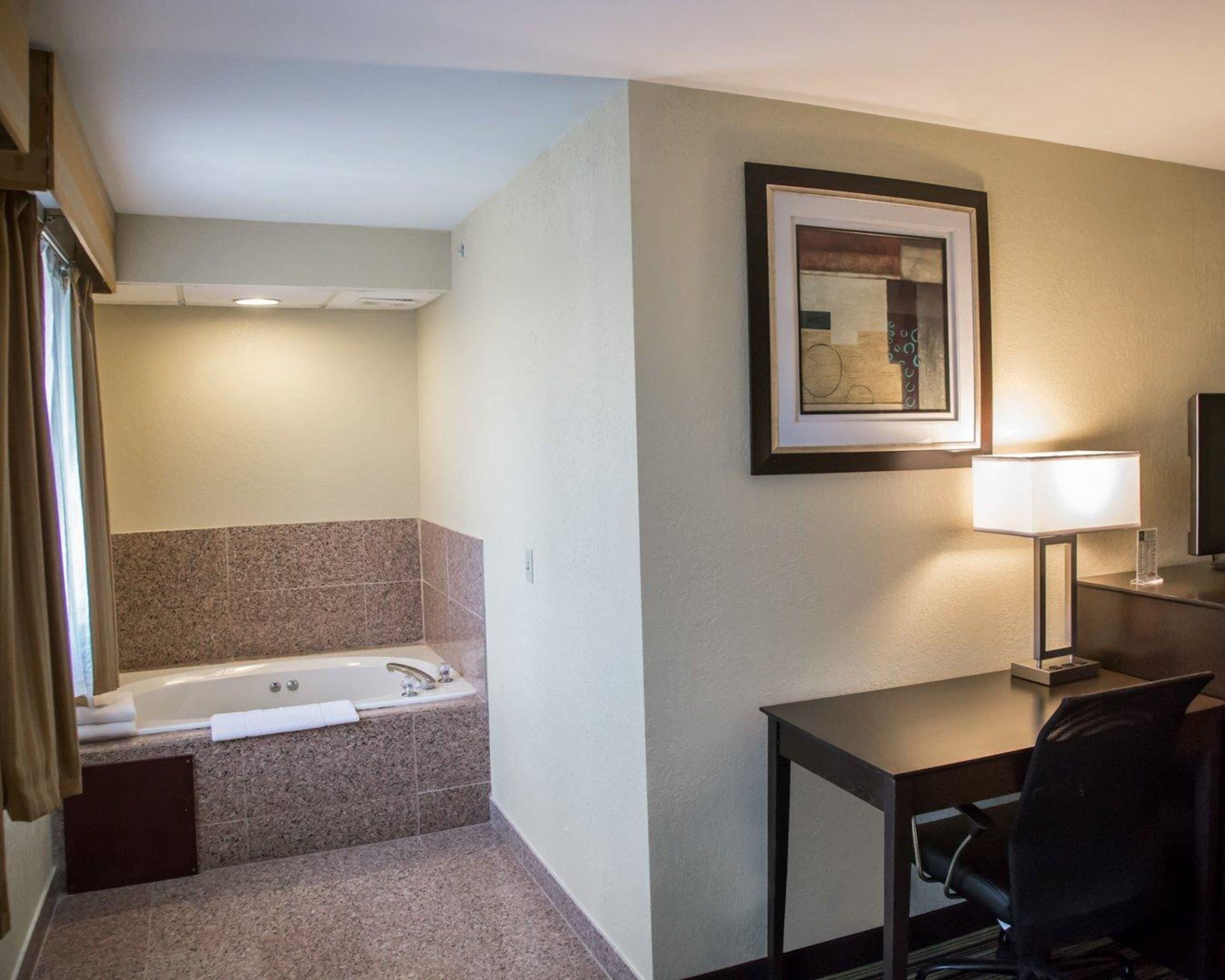 Une salle de bain d’hôtel avec une baignoire, un bureau avec une chaise, un tableau encadré au mur et une lampe.