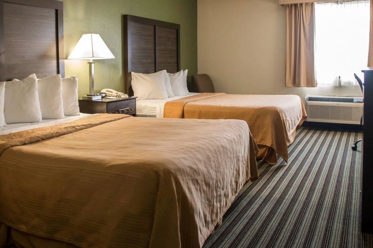 A hotel room with two beds, a nightstand, a lamp, an air conditioning unit, and a window with curtains.