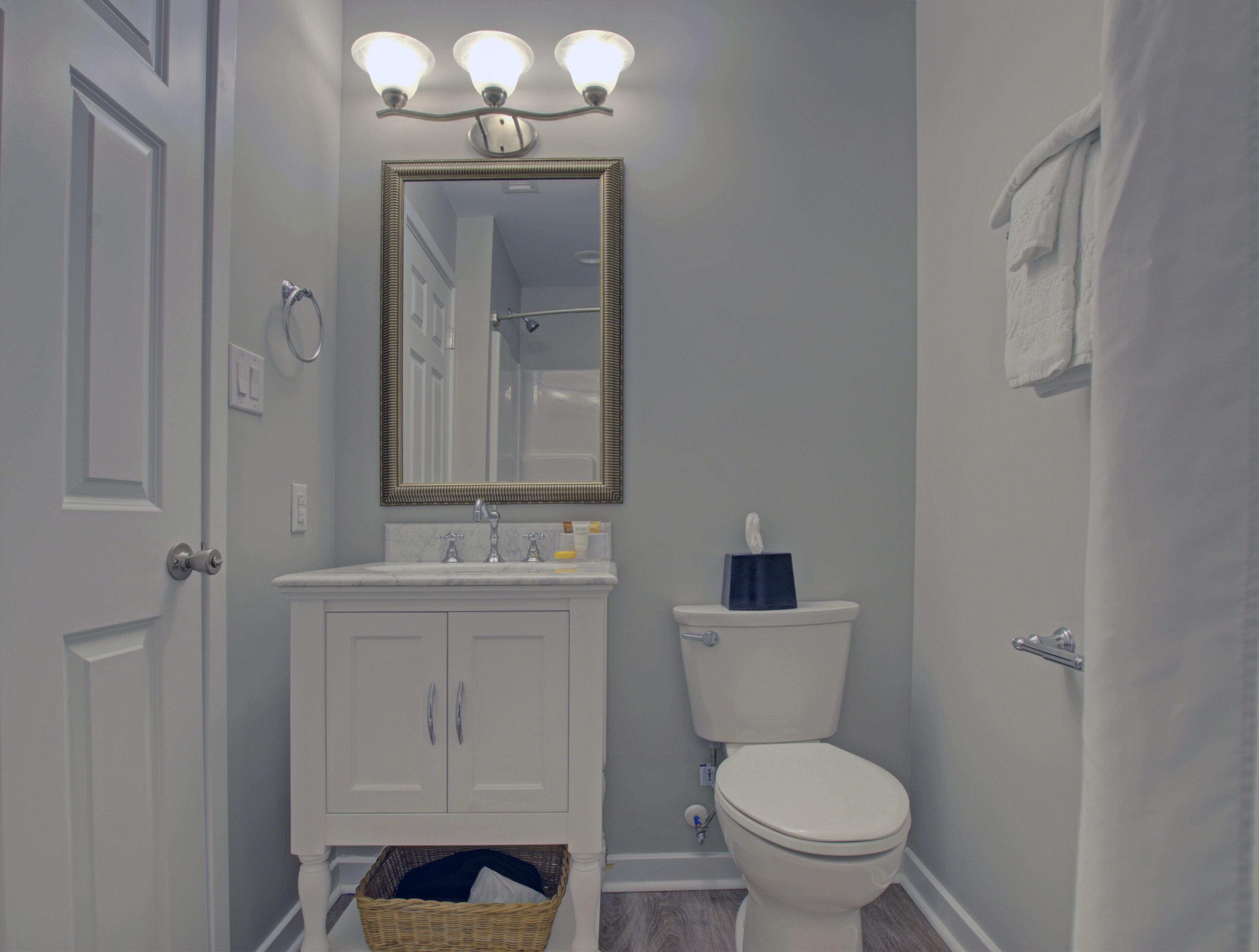 Ein Badezimmer mit weißem Waschtisch, einem Spiegel mit drei Lampen, einer Toilette und einer Duschvorhang.