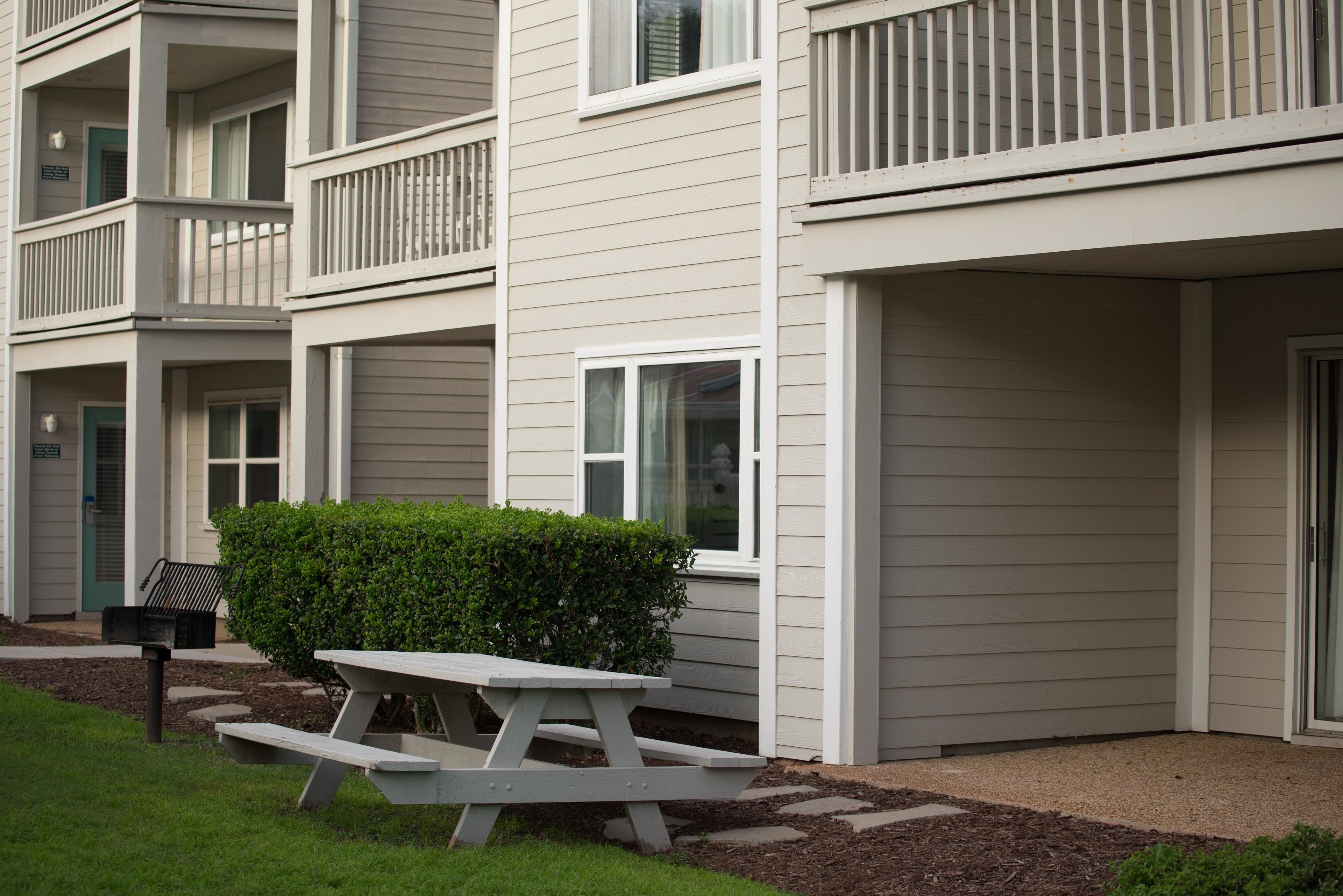 A multi-story residential building with a white exterior, balconies, and a picnic table in the front yard.