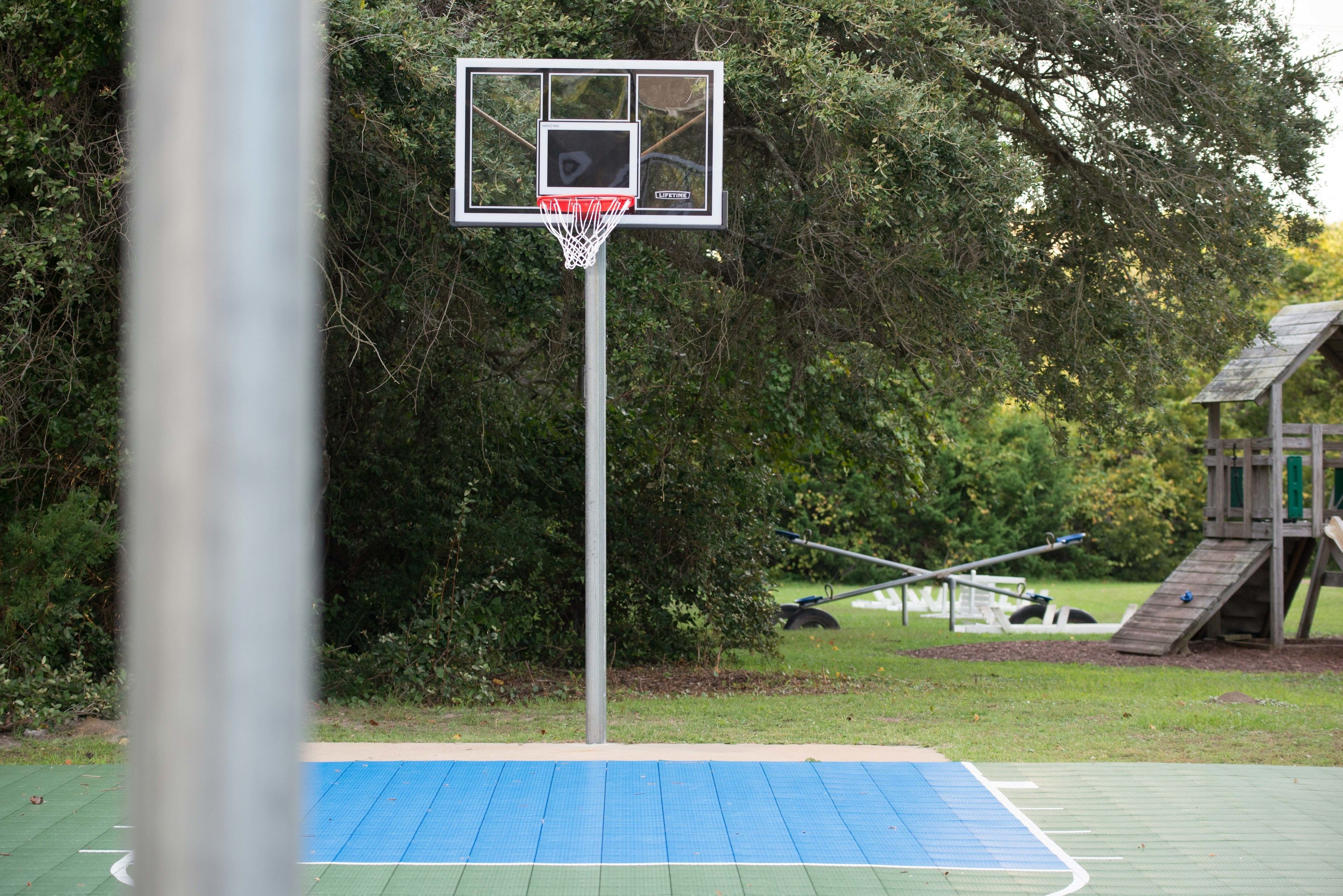 A basketball court with a hoop, surrounded by trees and a wooden play structure.
