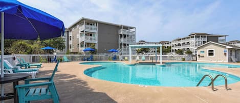 A swimming pool with lounge chairs and umbrellas, surrounded by buildings with balconies and a covered walkway.
