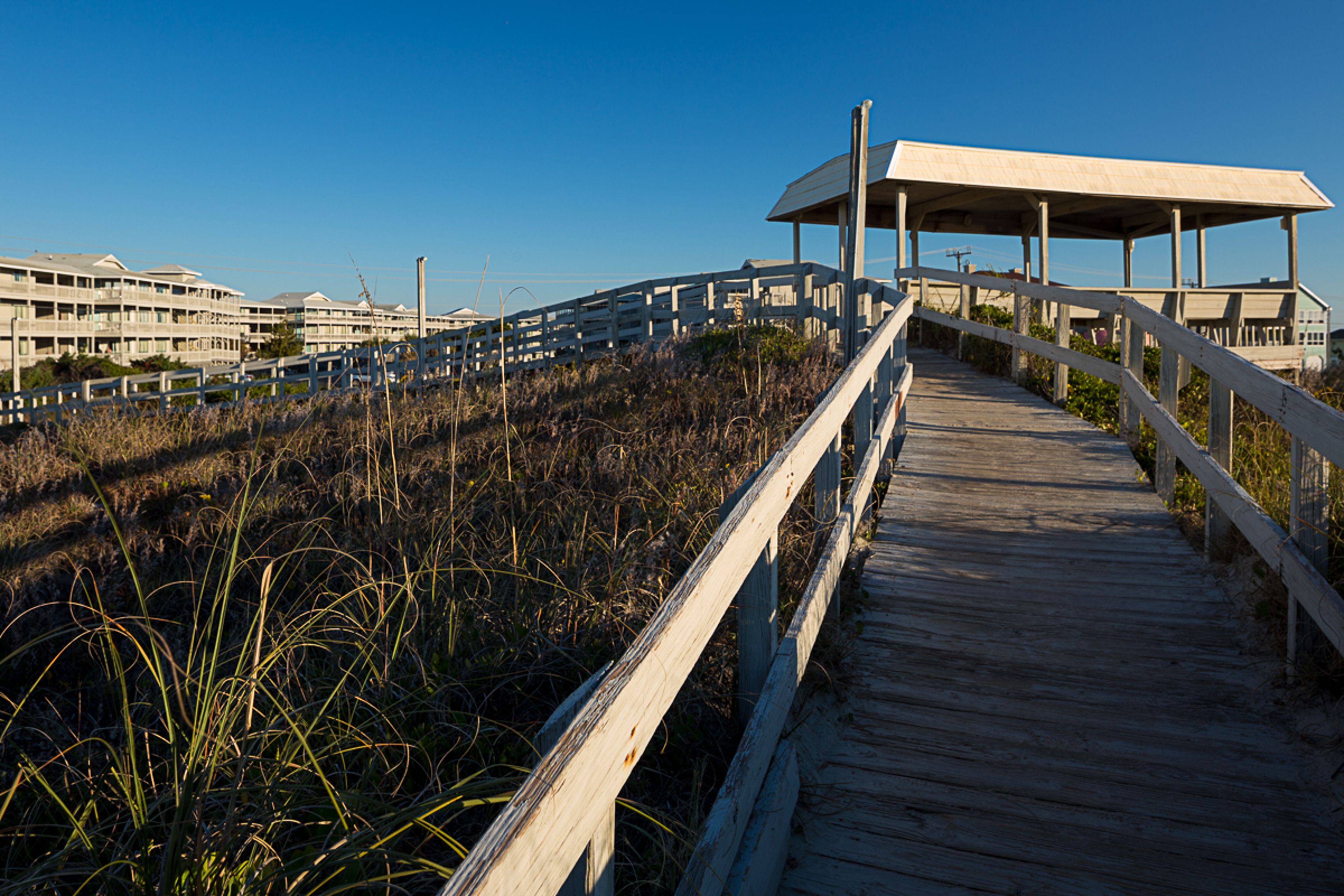 A wooden boardwalk leading to a covered pavilion with a view of buildings in the background.