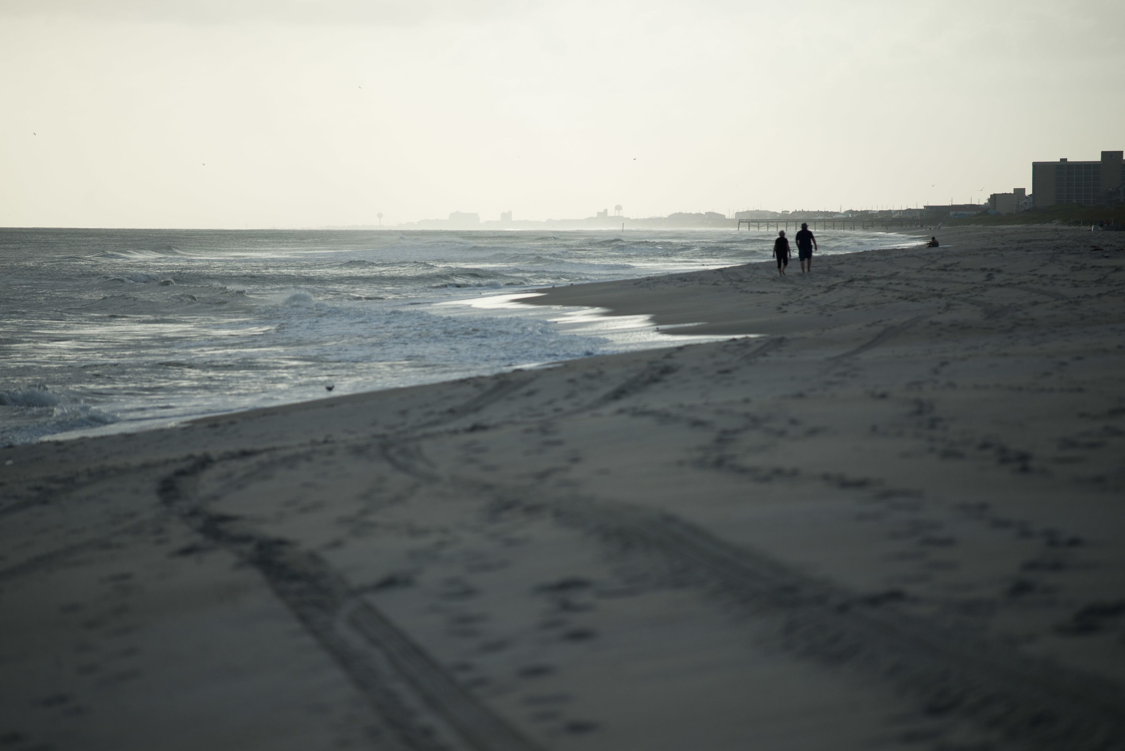 A beach with footprints leading to the water, a distant pier, and buildings in the background.