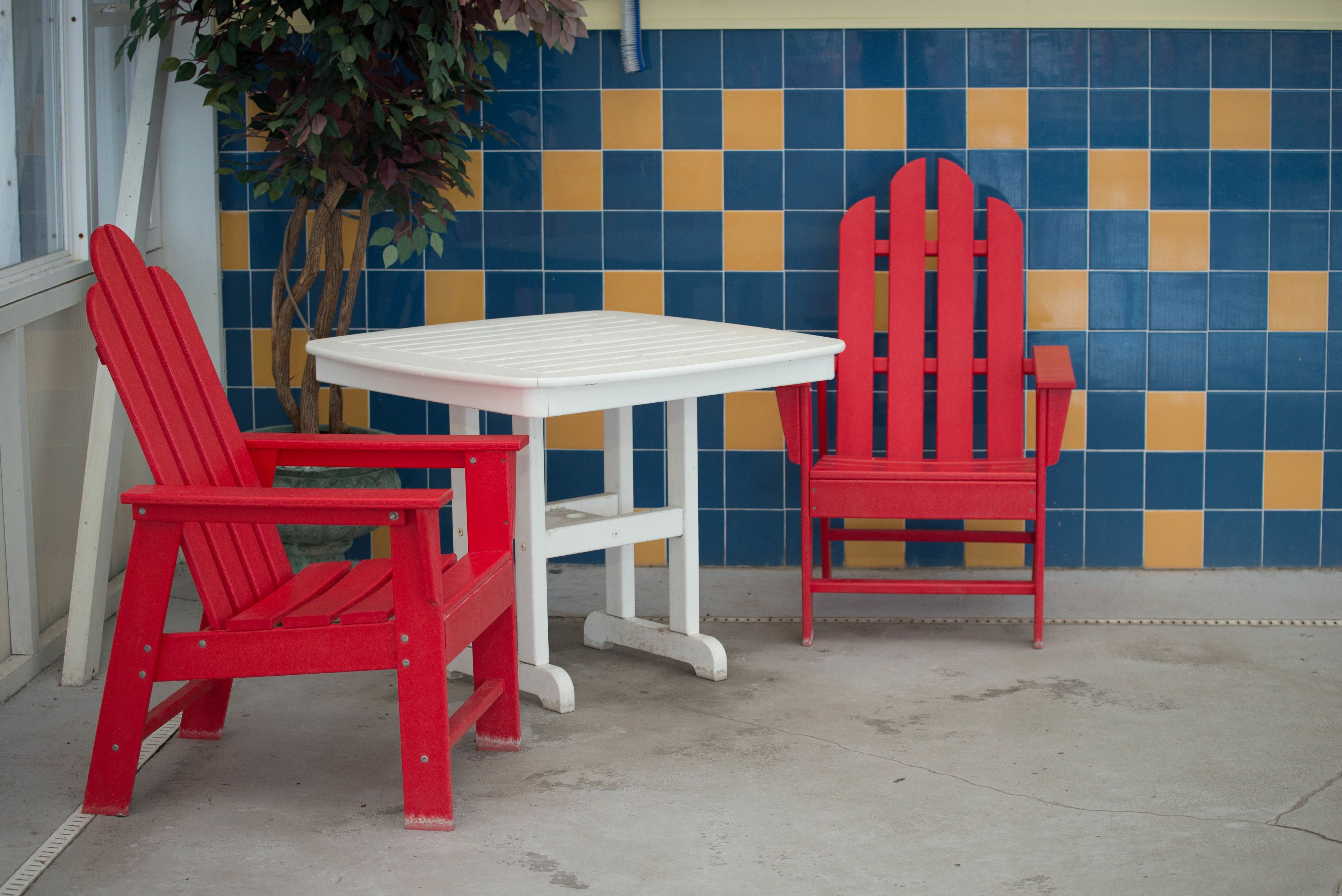 Two red Adirondack chairs and a white table against a blue and yellow tiled wall.