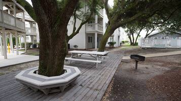 Une terrasse en bois, agrémentée de tables de pique-nique et de bancs, entourée d’arbres et bordée d’un bâtiment doté de balcons.