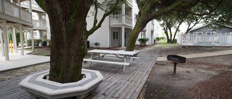 A wooden deck with picnic tables and benches, surrounded by trees and a building with balconies.