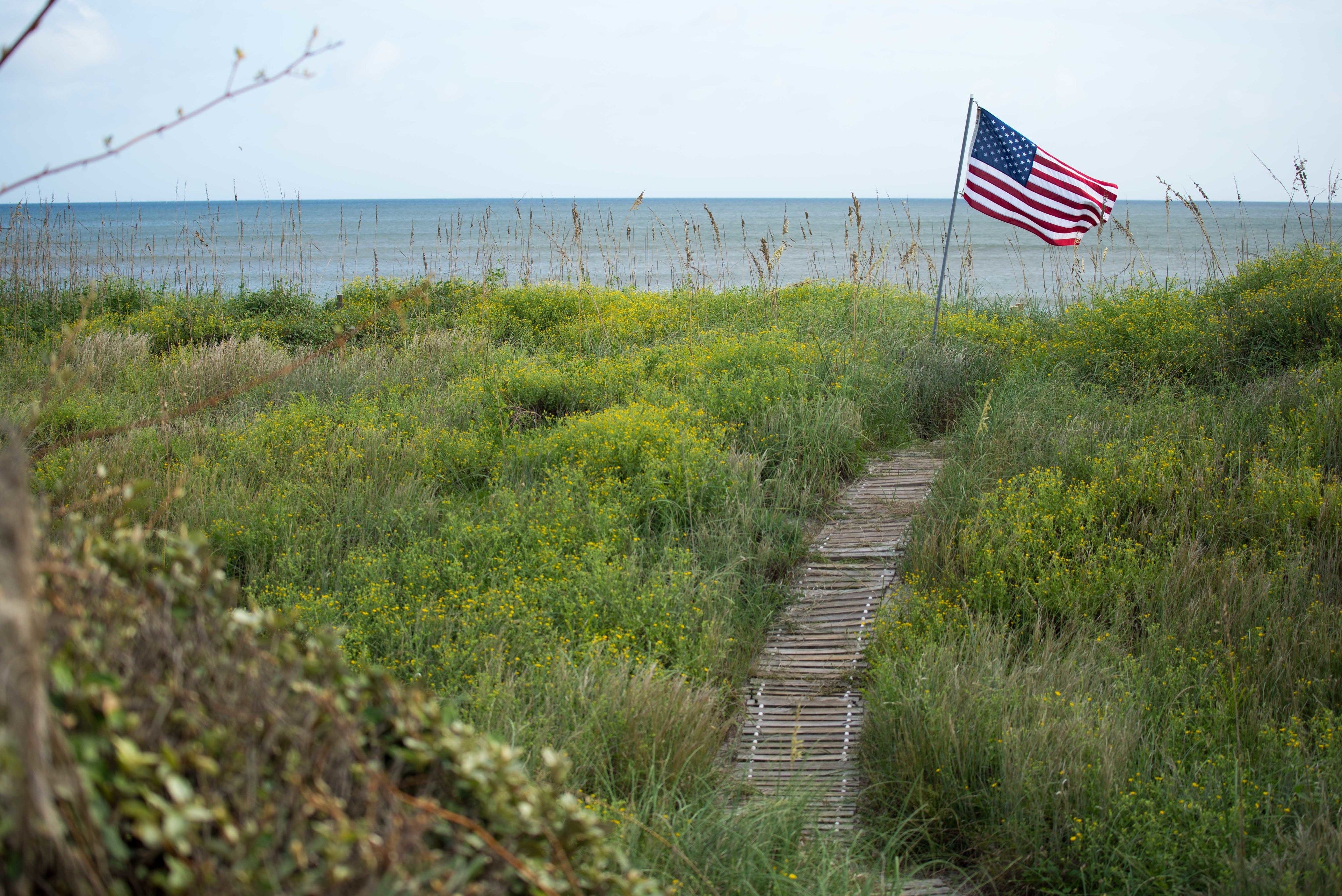 Ein hölzerner Steg führt zu einem Strand, eine amerikanische Flagge ist zu sehen.
