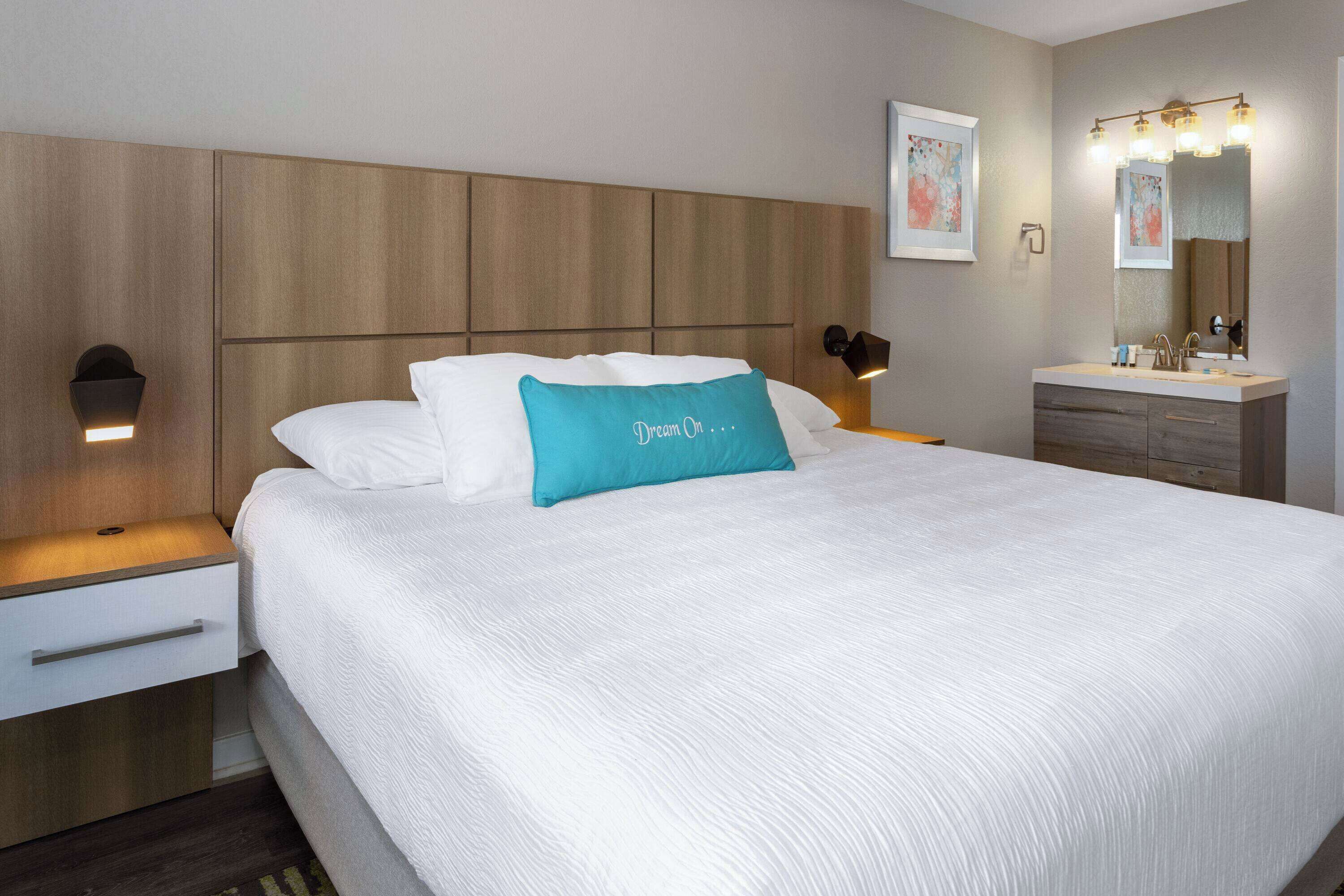 A neatly made bed with a white comforter and a teal pillow, a wooden headboard, bedside lamps, a nightstand with a drawer, and a bathroom with a vanity and mirror.